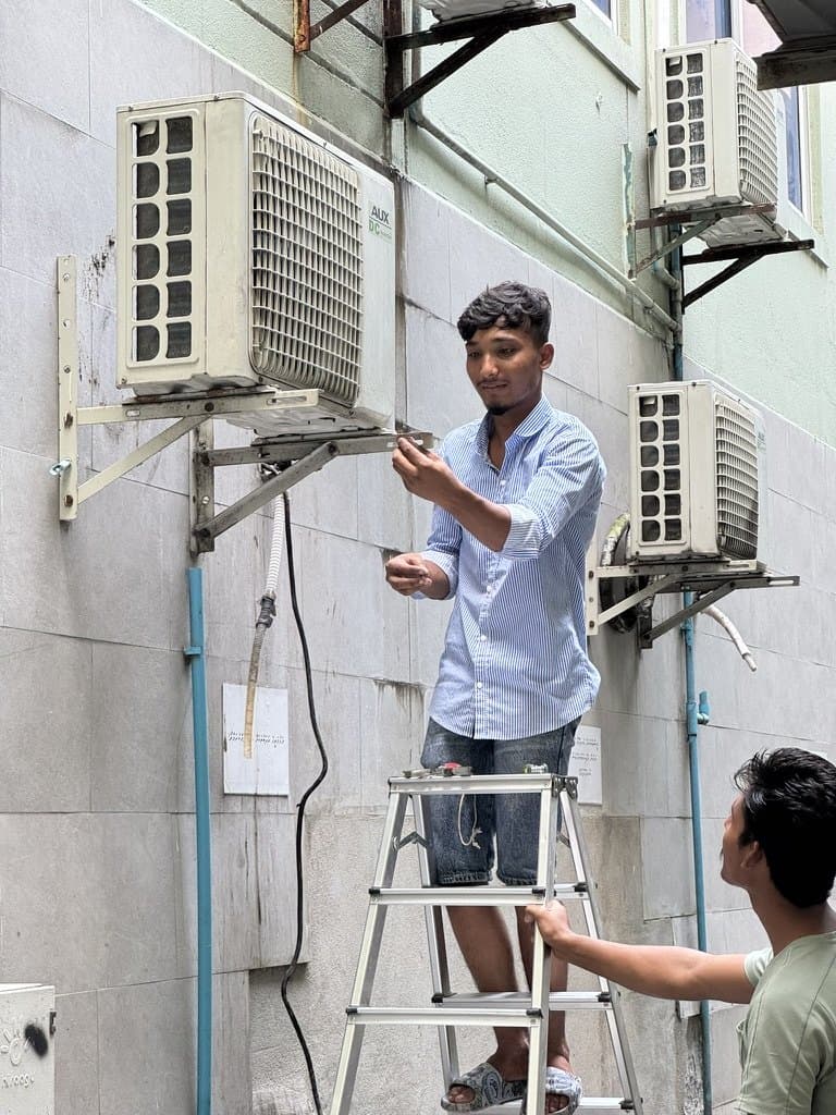 Rishthaa technician on a ladder at outdoor AC units mounted on a building wall, with a colleague steadying the ladder